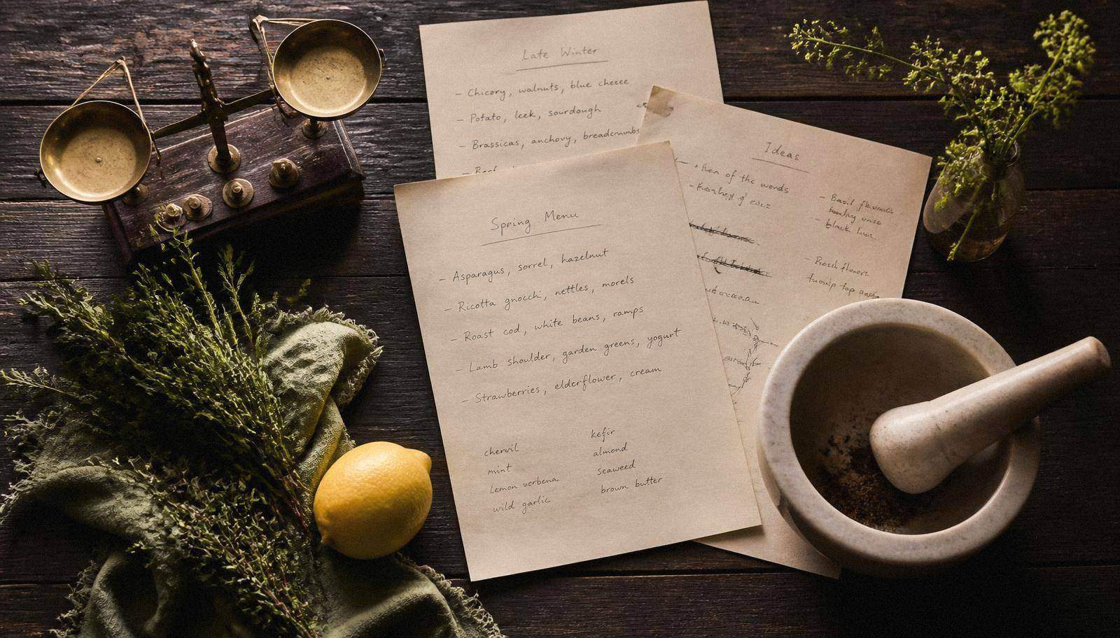 Handwritten menu drafts, a brass scale, herbs and a mortar and pestle on dark wood