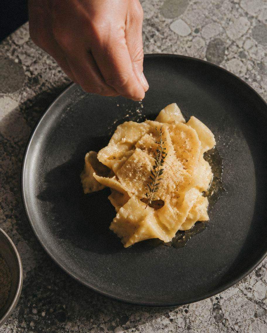 A chef's hand seasoning hand-torn pasta on a matte black stoneware plate