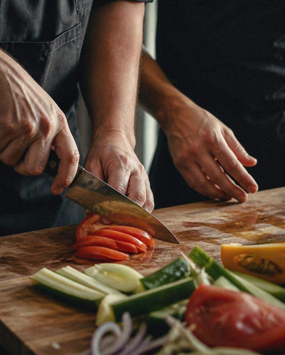 Two pairs of hands over a wooden board as a knife cut is demonstrated on fresh vegetables
